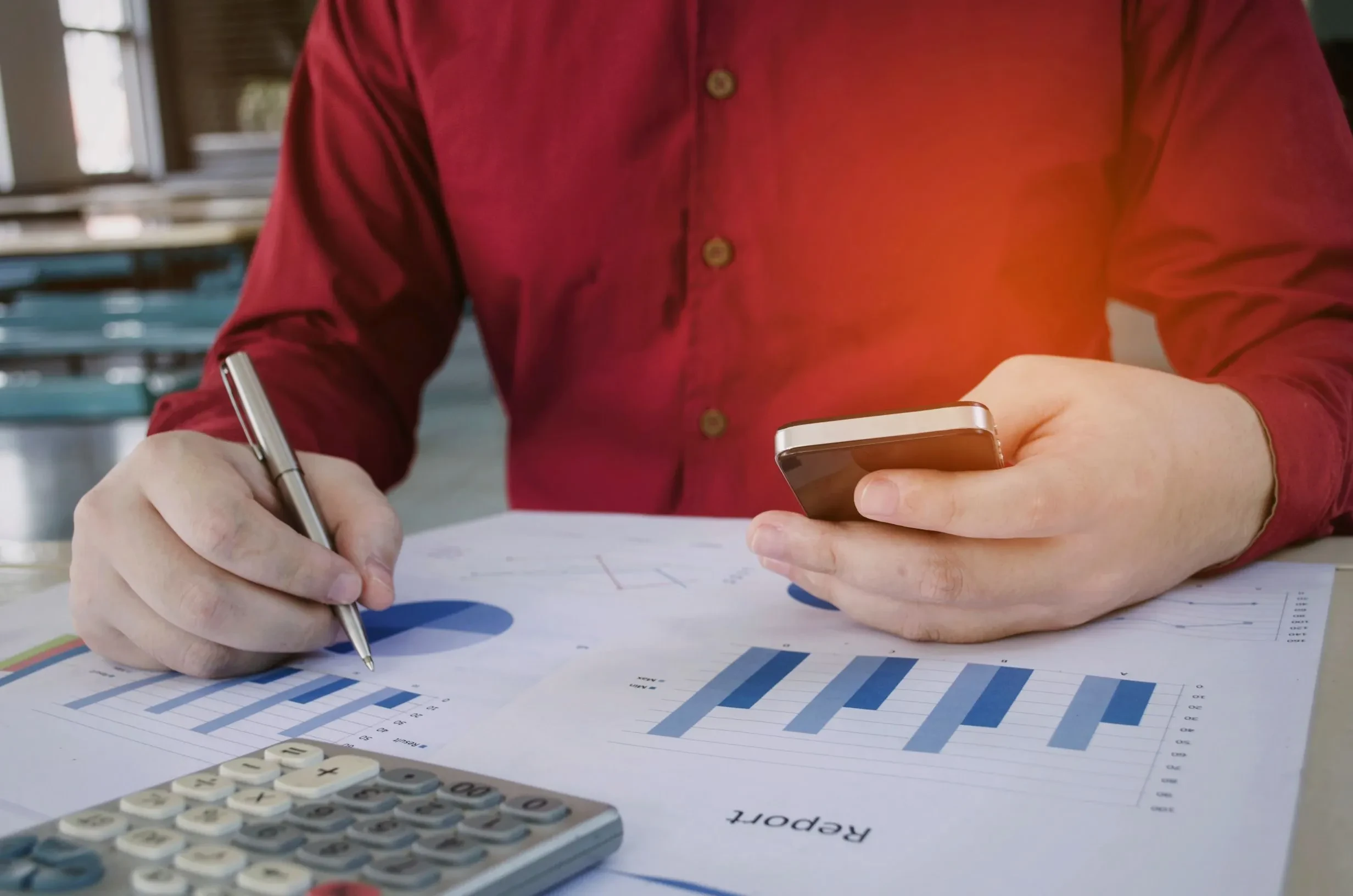 Person bei der Analyse eines Projektberichts mit Diagrammen, Smartphone und Taschenrechner – Symbolbild für Projektcontrolling und datenbasierte Entscheidungsfindung im Multiprojektmanagement