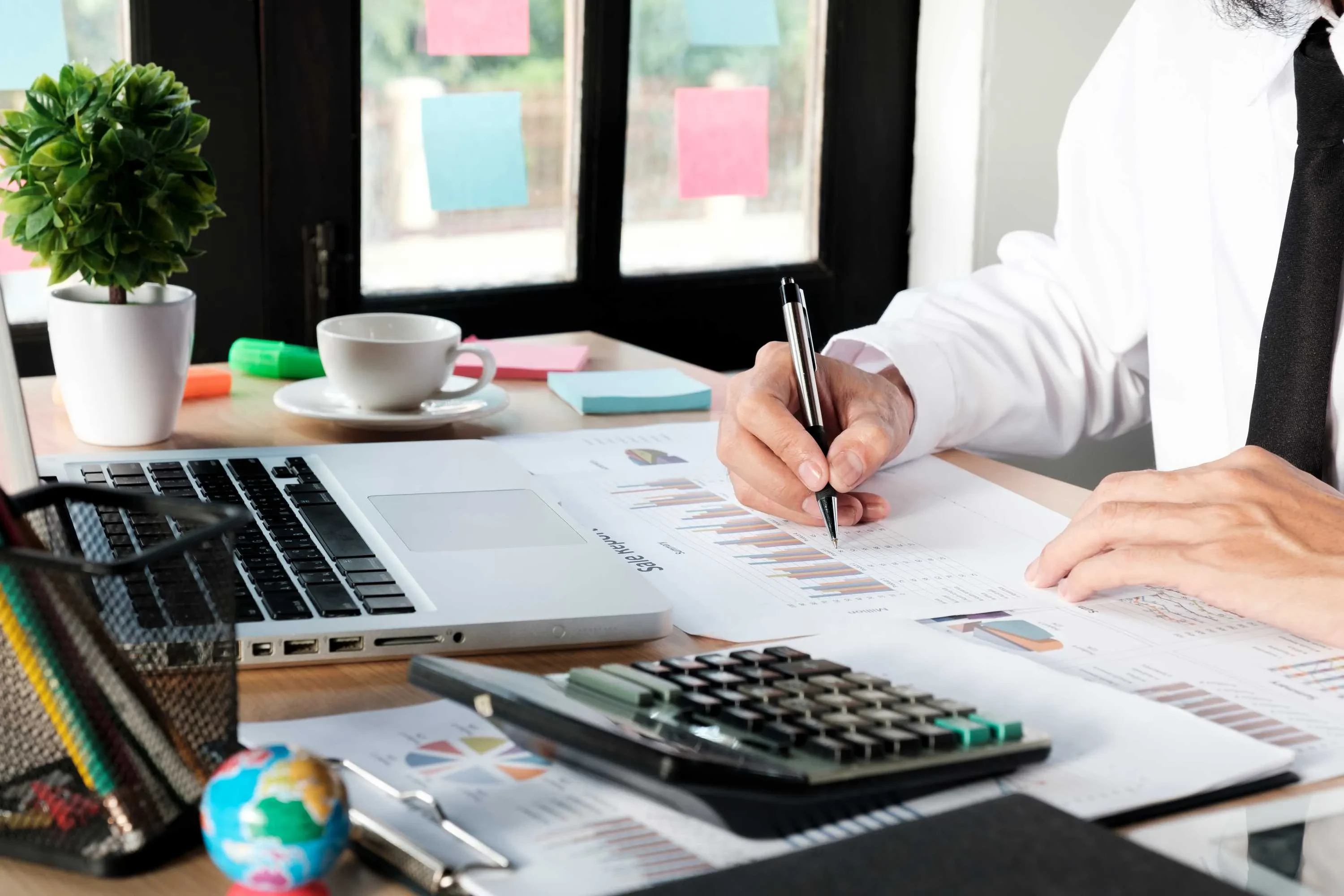 Person bei der Analyse von Diagrammen und Berichten an einem Schreibtisch mit Laptop, Kaffee und Globus – visuelles Motiv für strategisches Projektmanagement und datenbasierte Planung