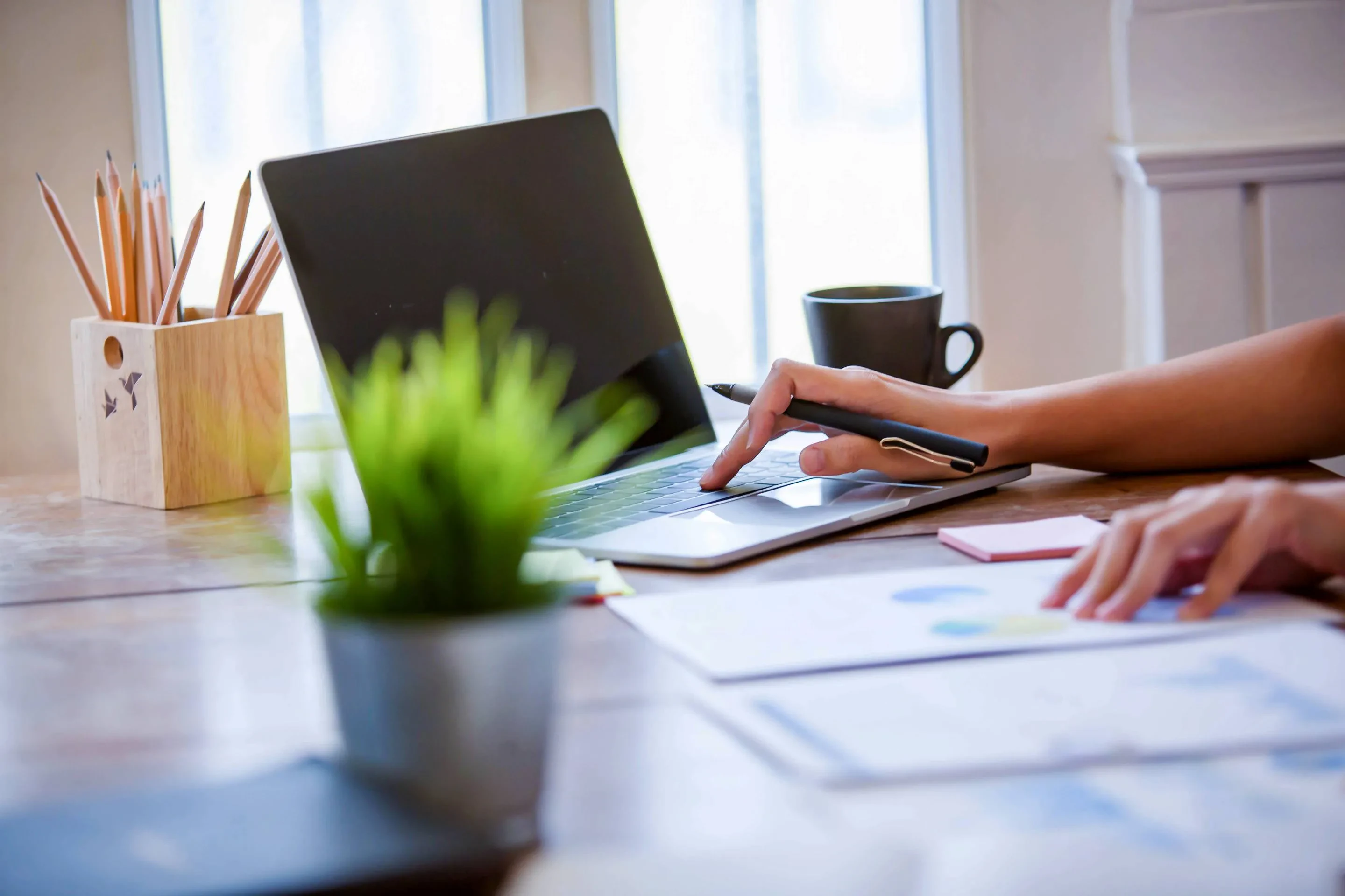Person zeigt mit einem Stift auf den Laptopbildschirm an einem Schreibtisch mit Kaffeetasse, Pflanze und Charts – Symbol für digitale Projektarbeit und datenbasierte Planung