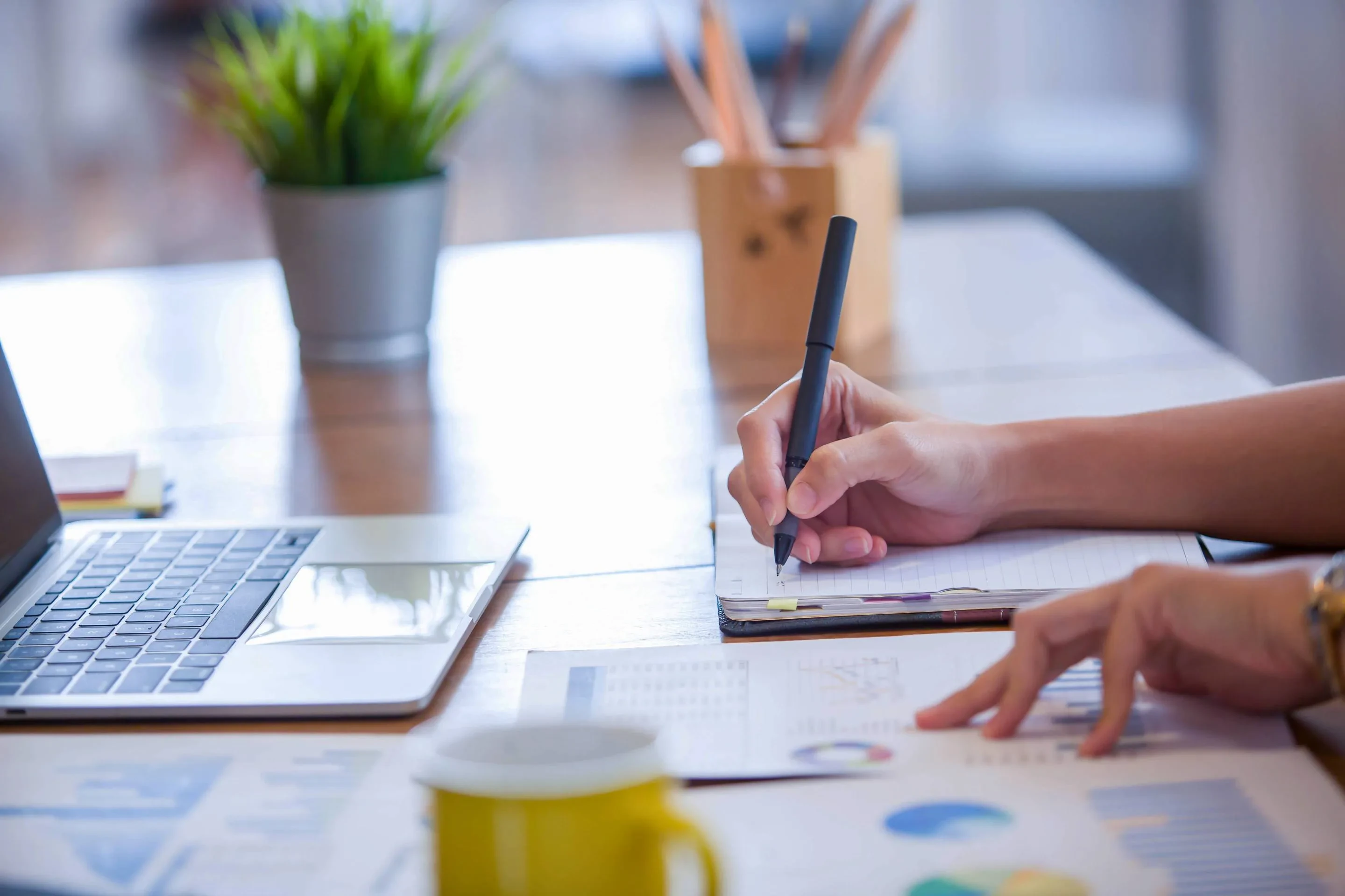 Person schreibt in ein Notizbuch und zeigt auf gedruckte Diagramme – Symbol für datenbasierte Planung und Projektanalyse im modernen Arbeitsumfeld