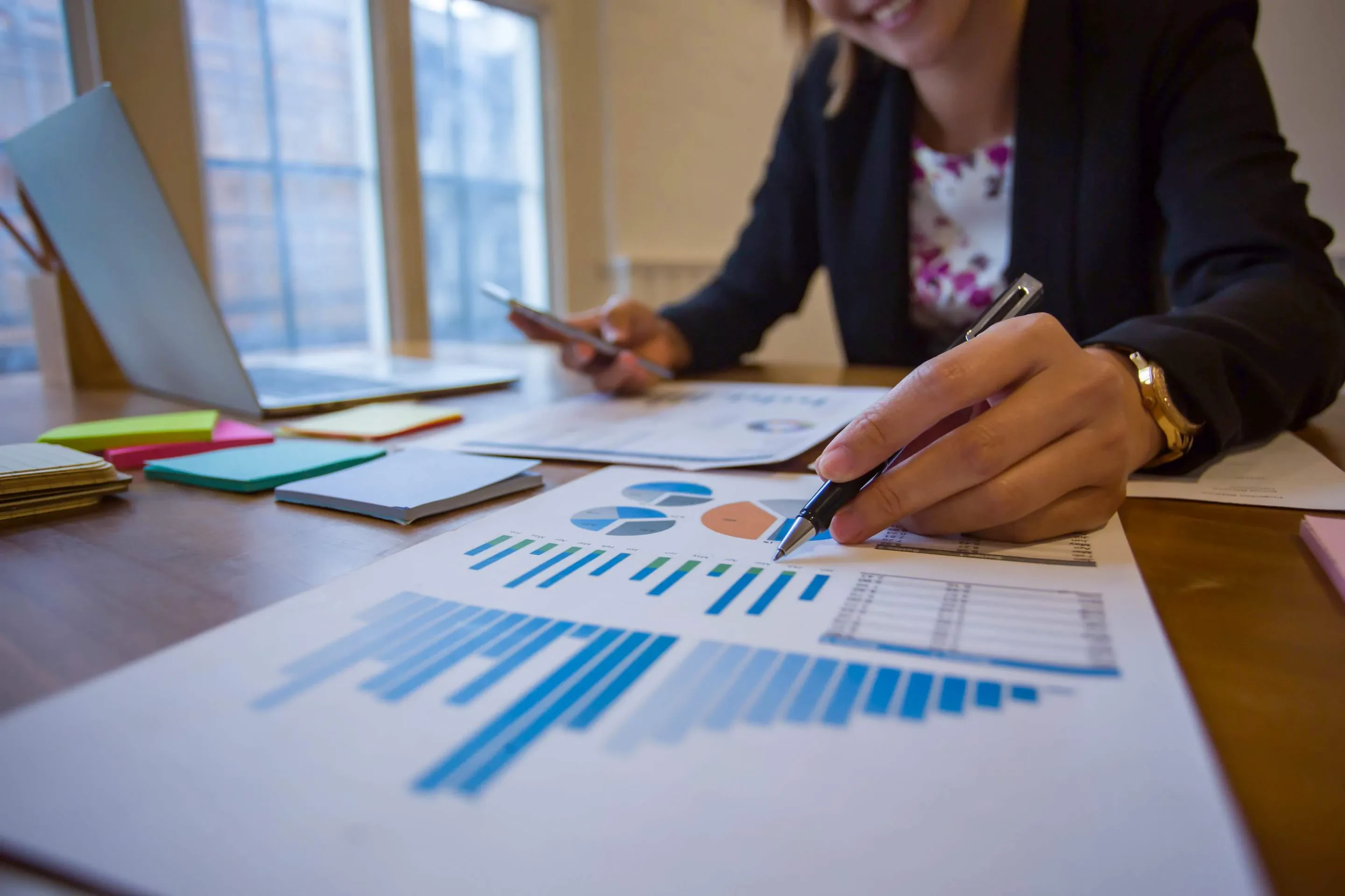 Person zeigt mit Stift auf Balkendiagramm und hält Smartphone – Symbol für visuelle Projektanalyse und Reporting im Büroalltag