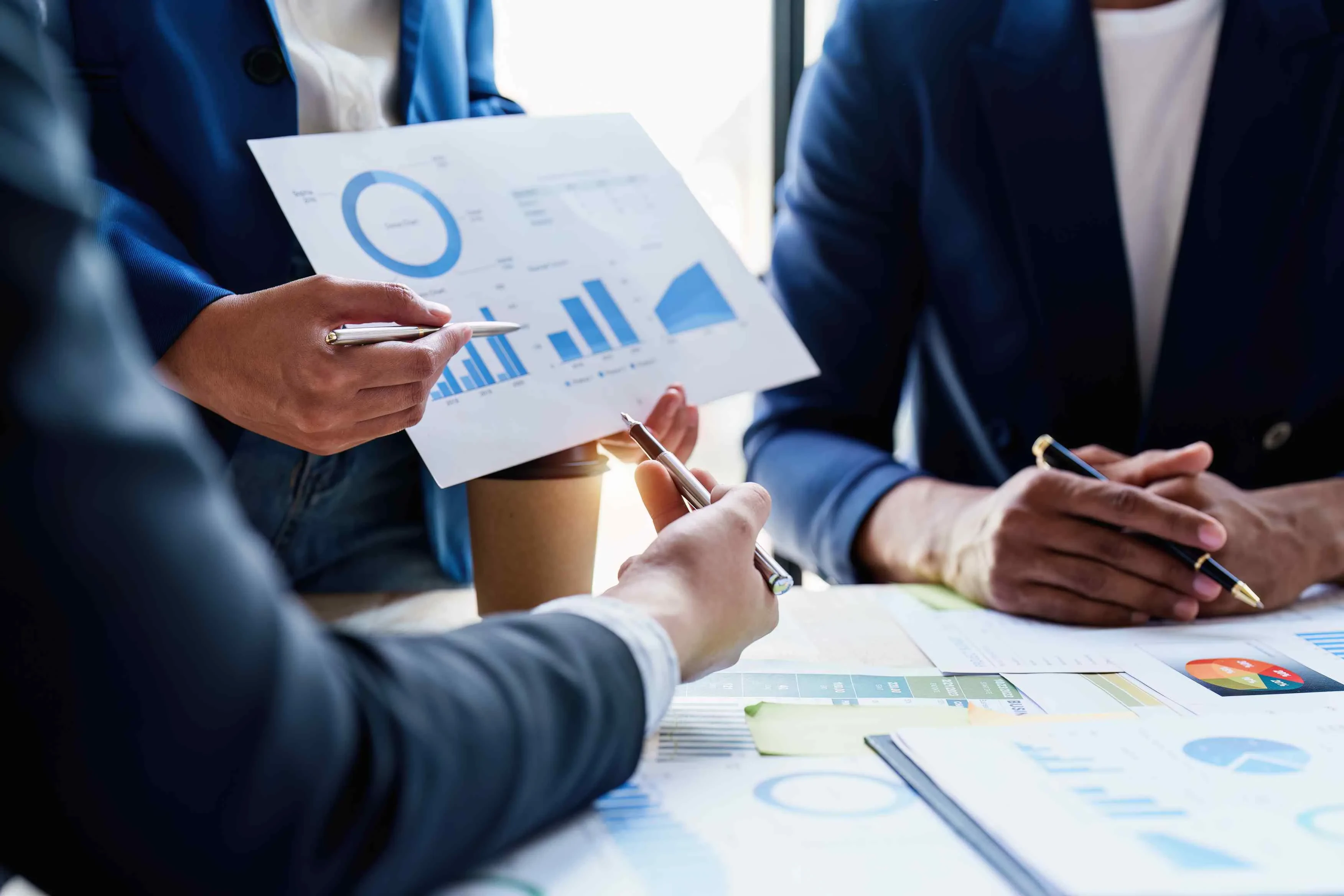 Drei Personen mit Charts und Kaffeetasse im Gespräch – Symbol für datenbasierte Teamarbeit und strategische Projektplanung.